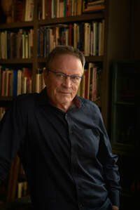 A man in a blue shirt and glasses is standing in front of a bookshelf.