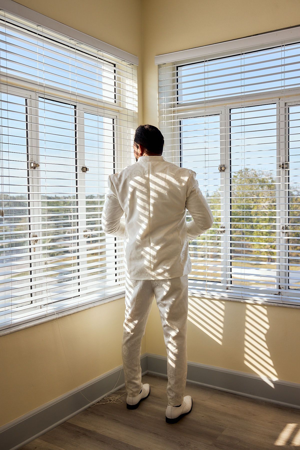 A man in a white suit is standing in front of a window with blinds.