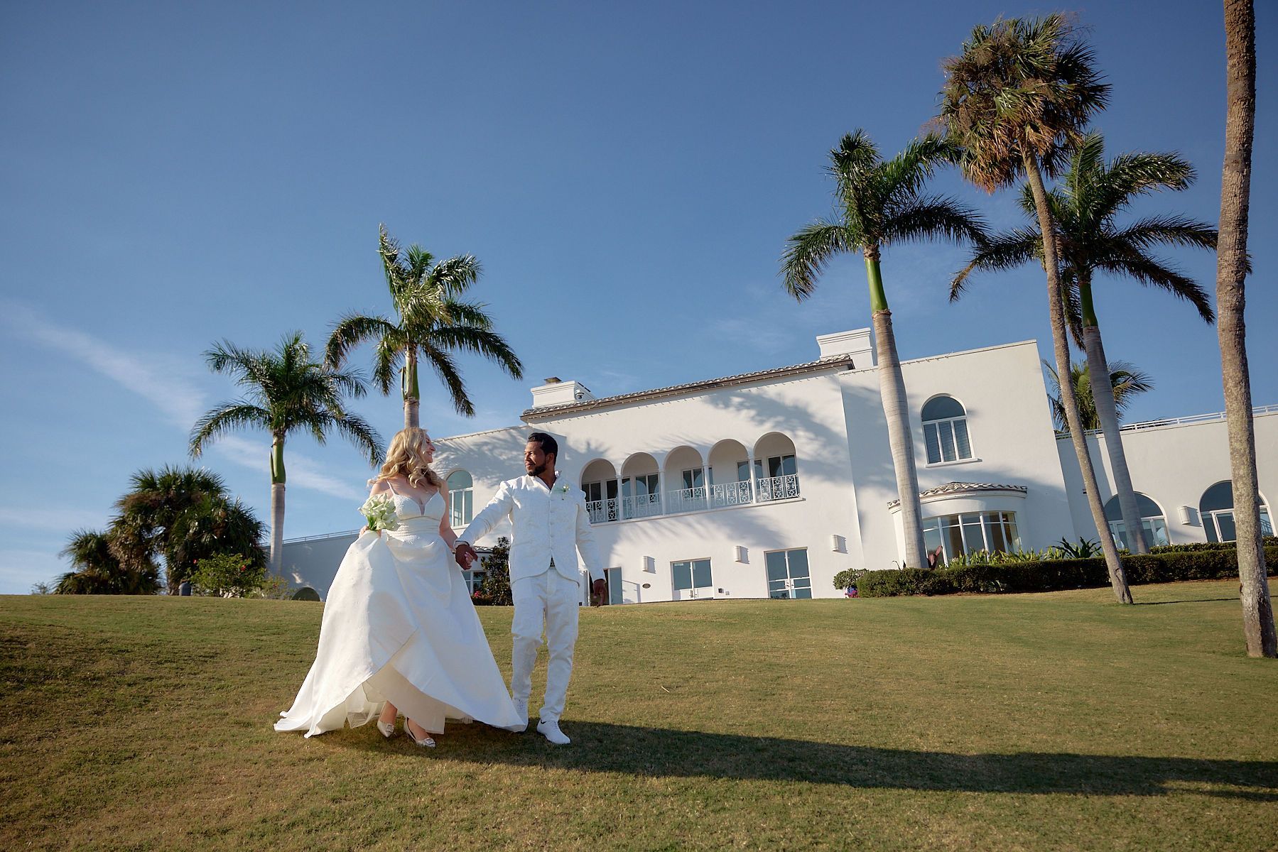 A bride and groom are walking in front of a white building holding hands.