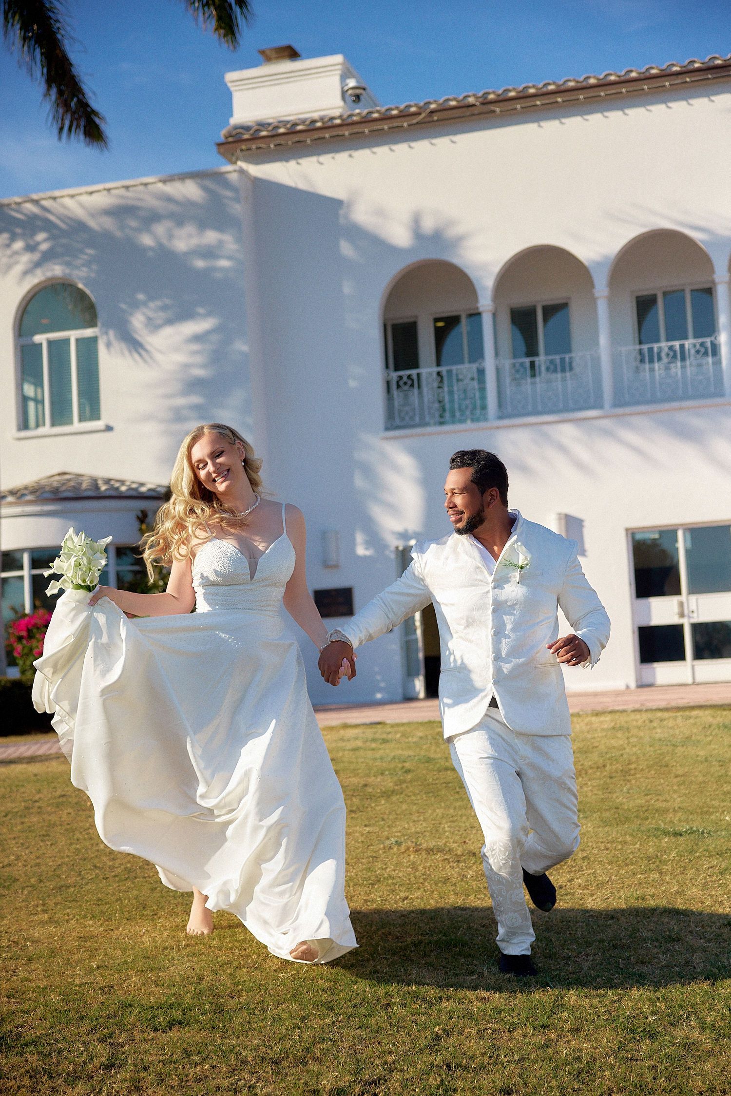 A bride and groom are running in front of a white building holding hands.