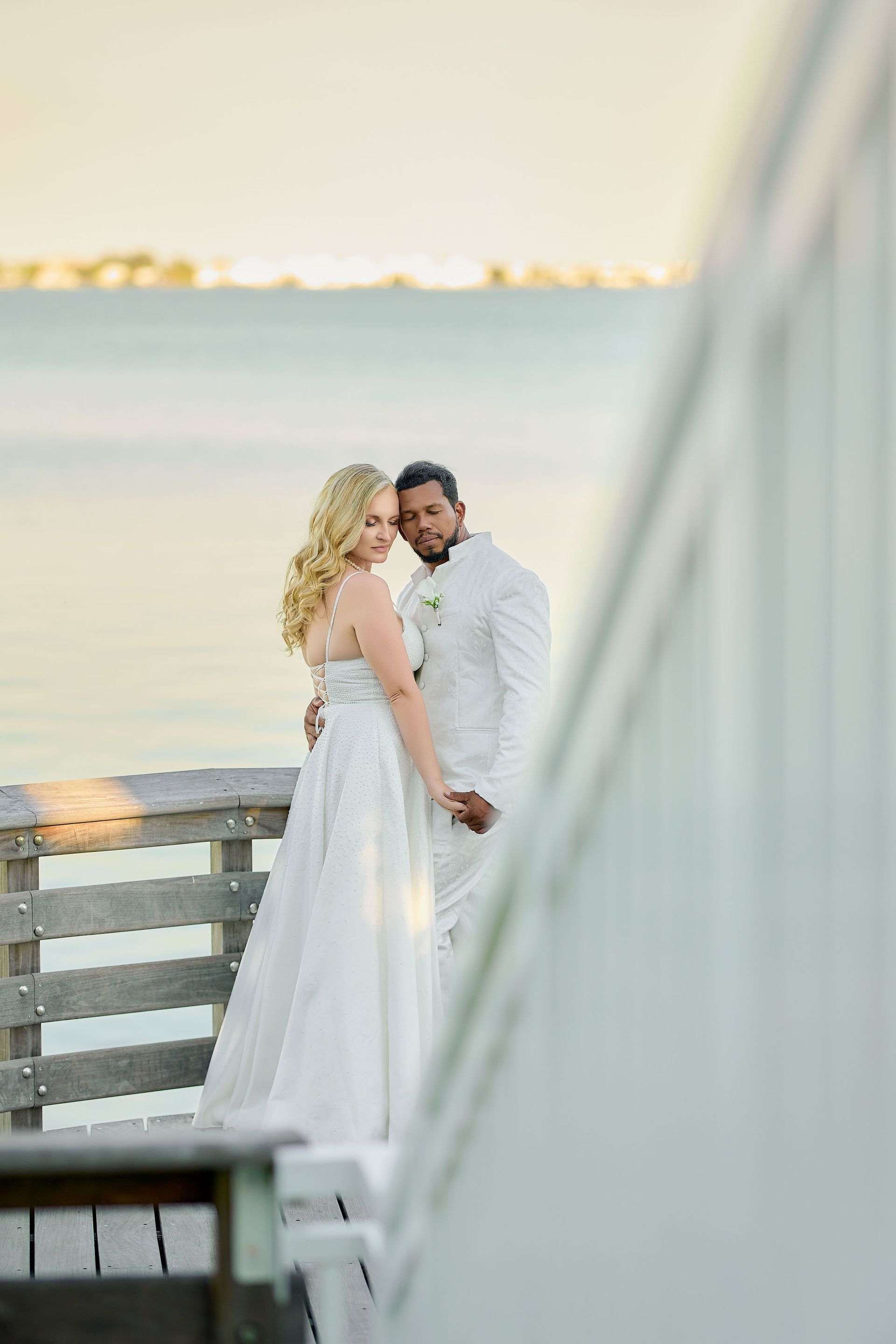 A bride and groom are posing for a picture on a dock overlooking the ocean.