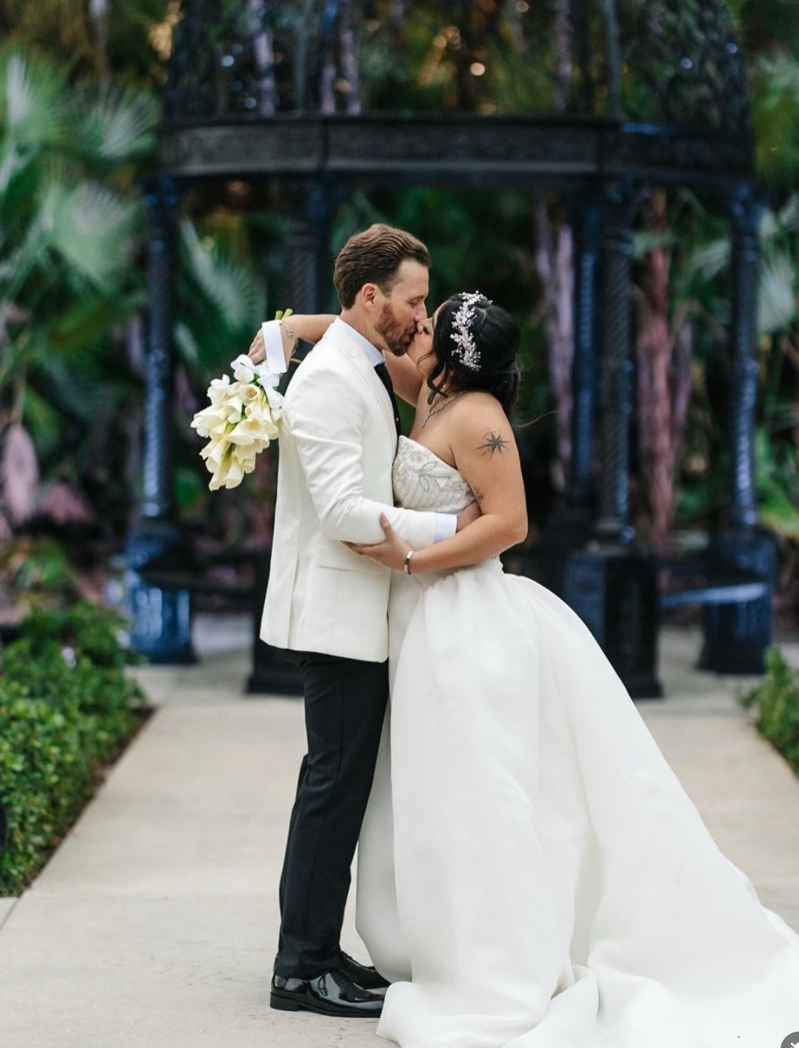 Couple kissing, embracing; the bride in a white gown, holding a bouquet; groom in a white jacket; in front of a gazebo.