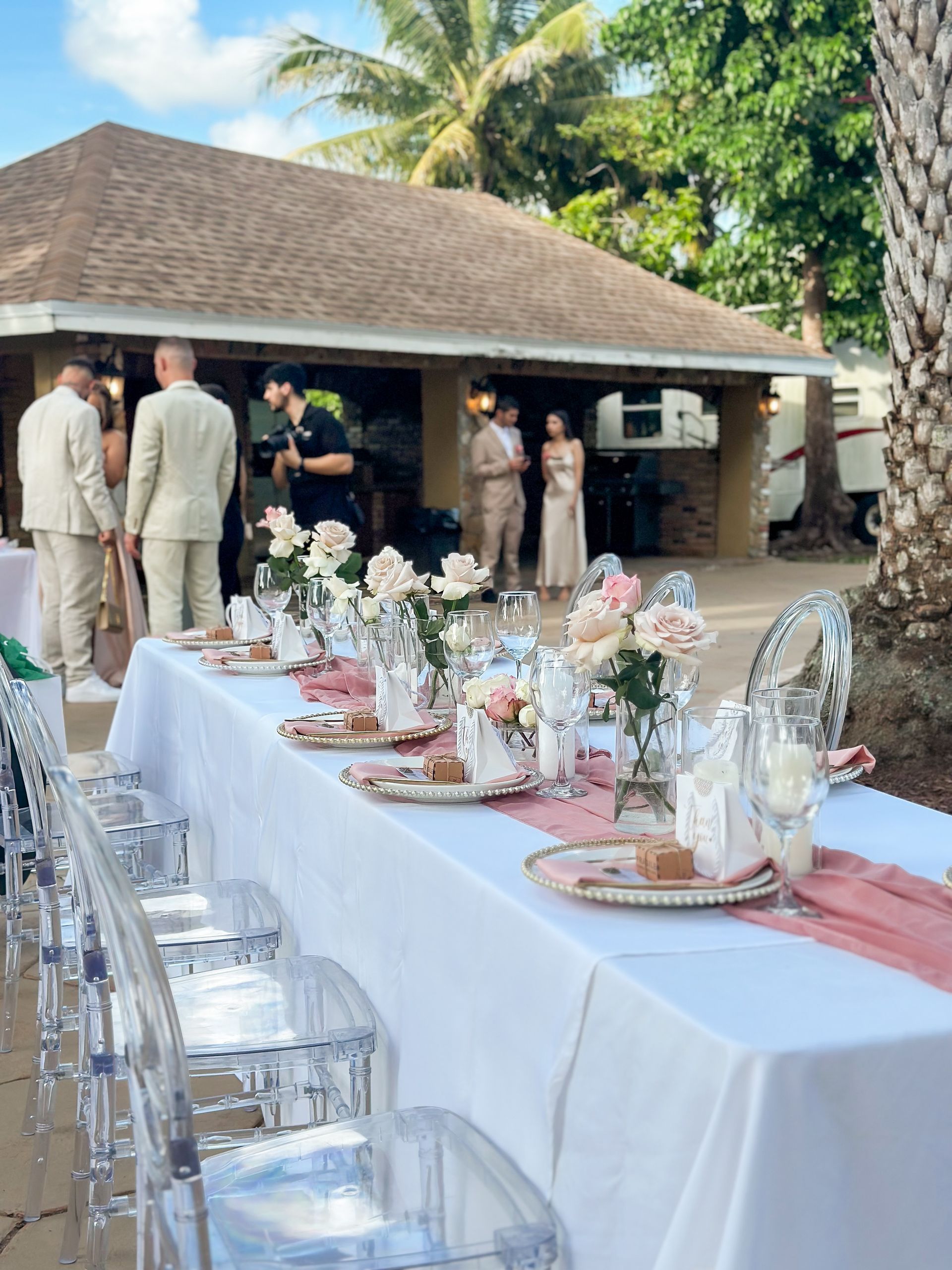 A long table with a white table cloth and clear chairs is set for a wedding reception.
