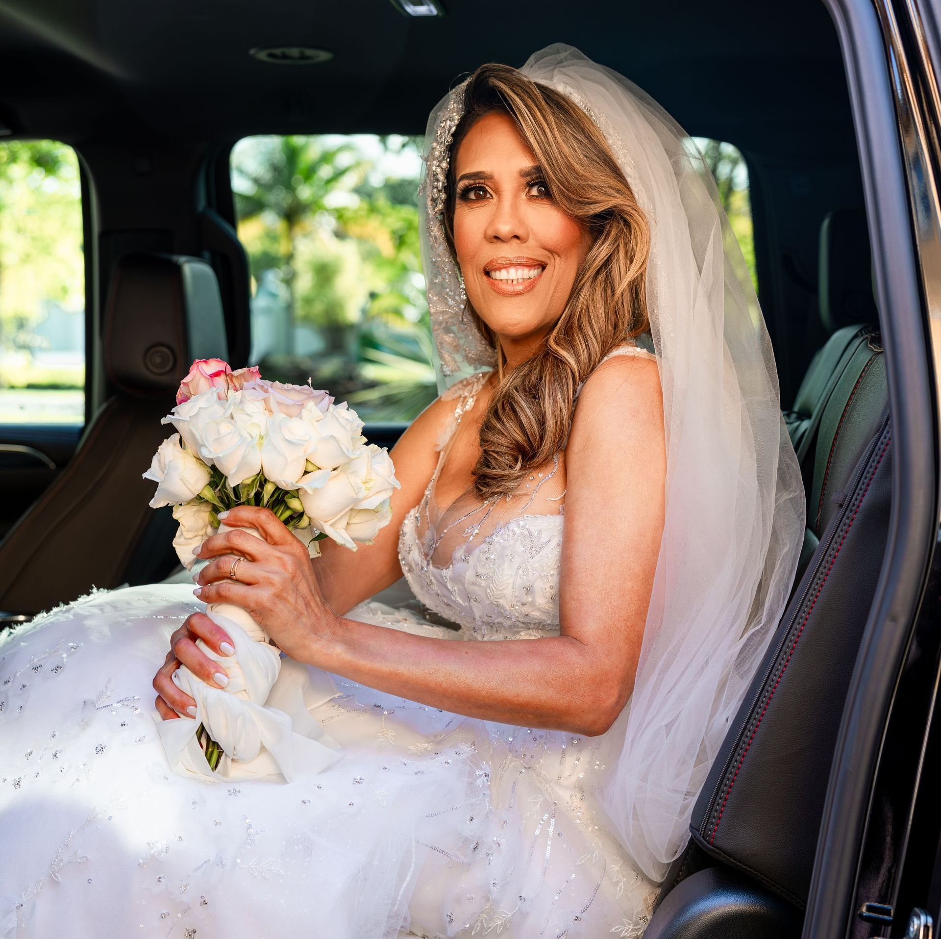 A bride is sitting in the back seat of a car holding a bouquet of flowers