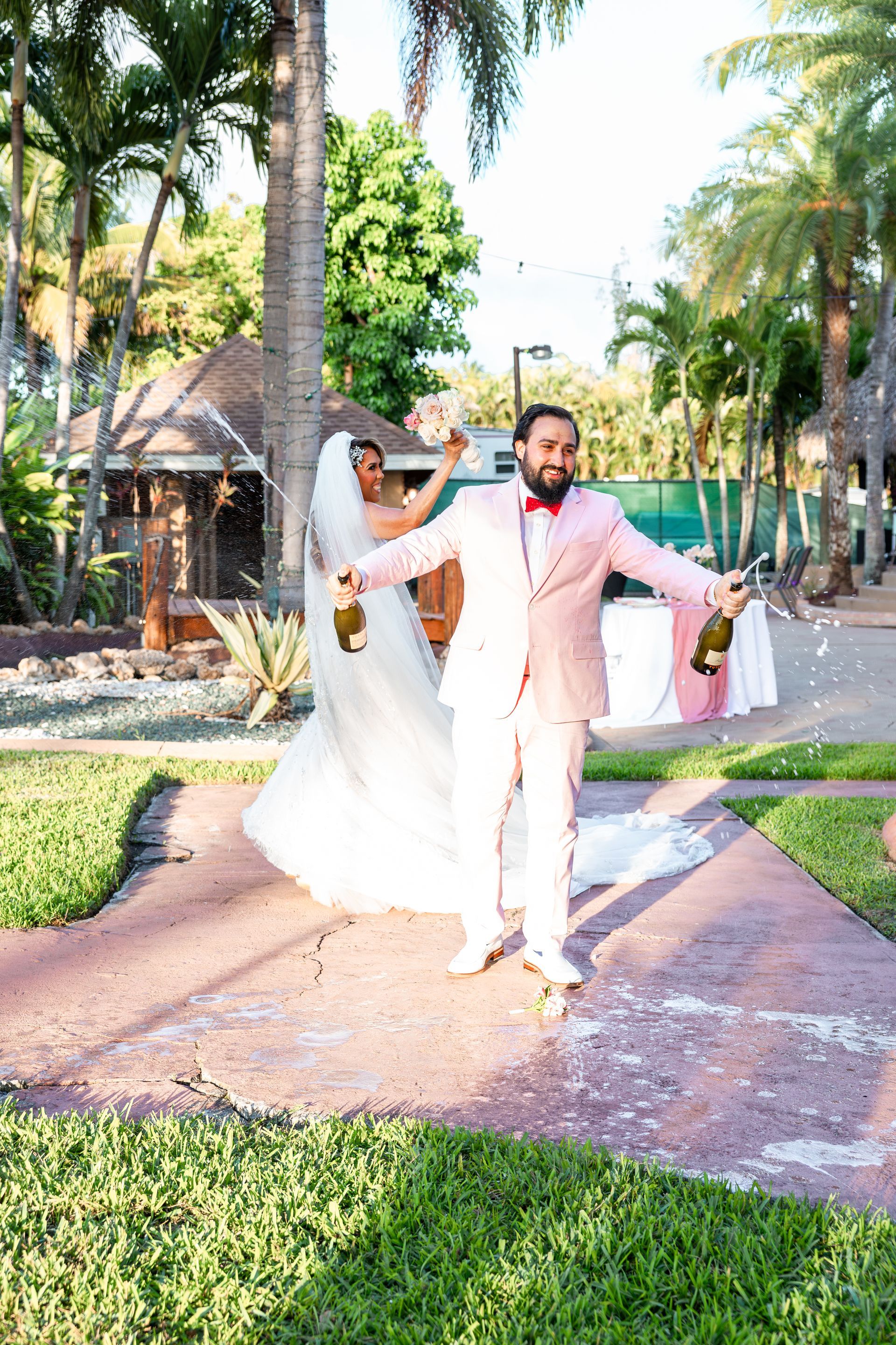A bride and groom are walking down a sidewalk holding champagne bottles.