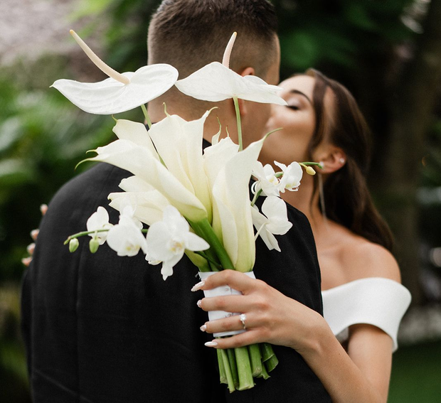 Bride kisses groom, holding white bouquet, both in formal wear, outdoors.