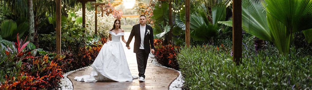 A newlywed couple walks hand-in-hand down a garden path. The bride wears a white gown, and the groom a suit.
