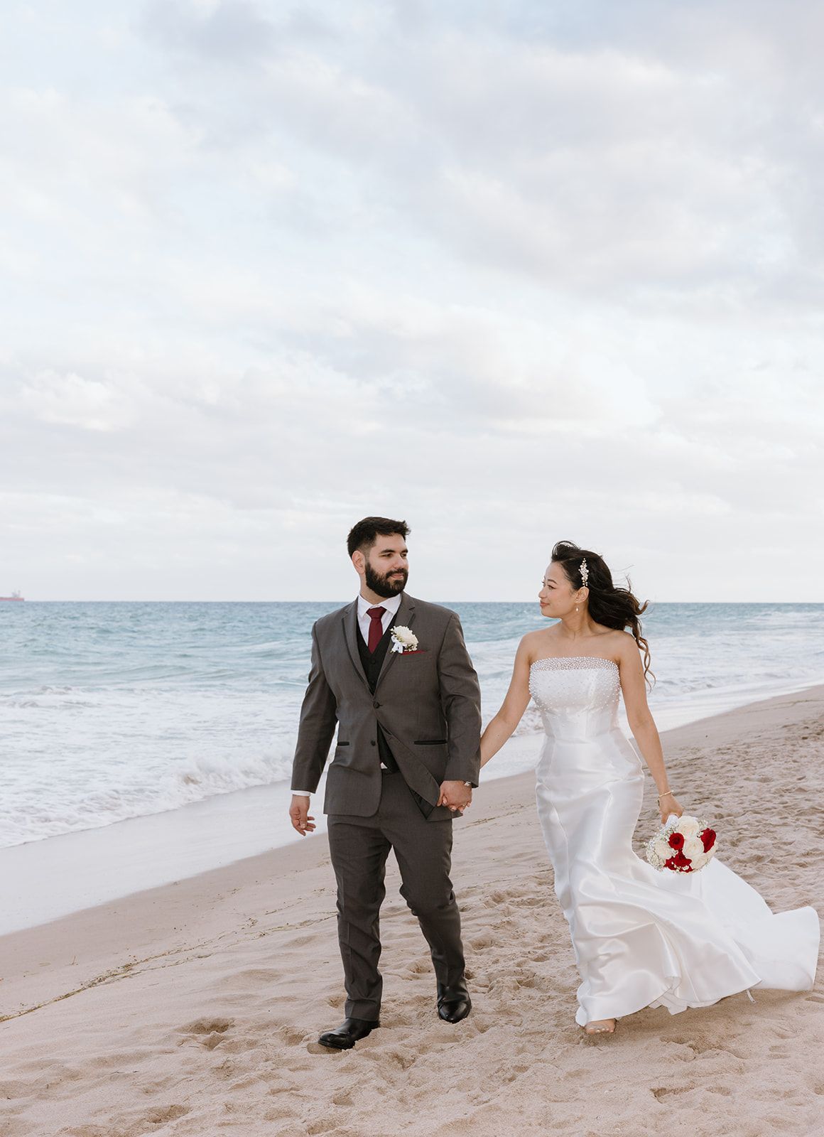 A bride and groom are walking on the beach holding hands.