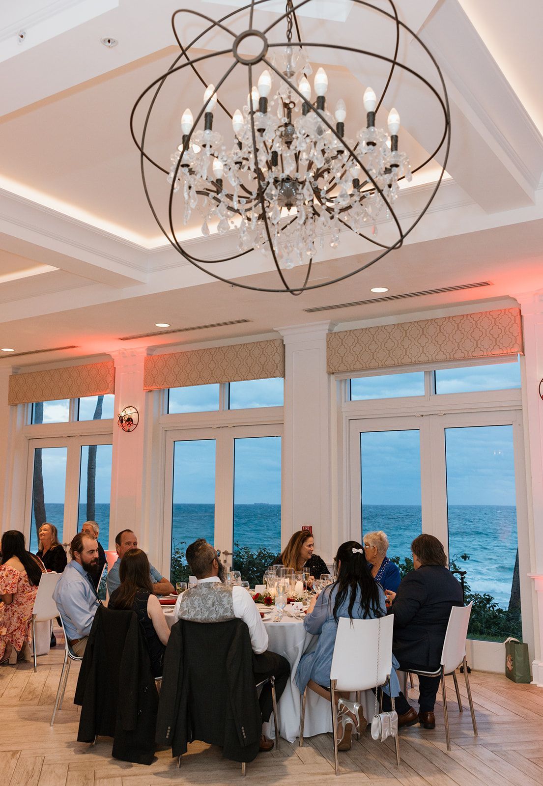 A group of people are sitting at tables in a room with a chandelier hanging from the ceiling.