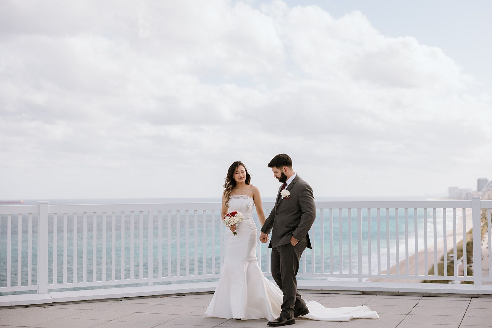 A bride and groom are walking on a balcony overlooking the ocean.