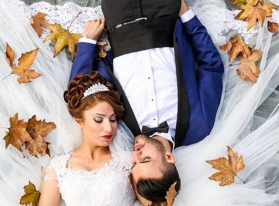 A bride and groom are laying on their backs surrounded by leaves.