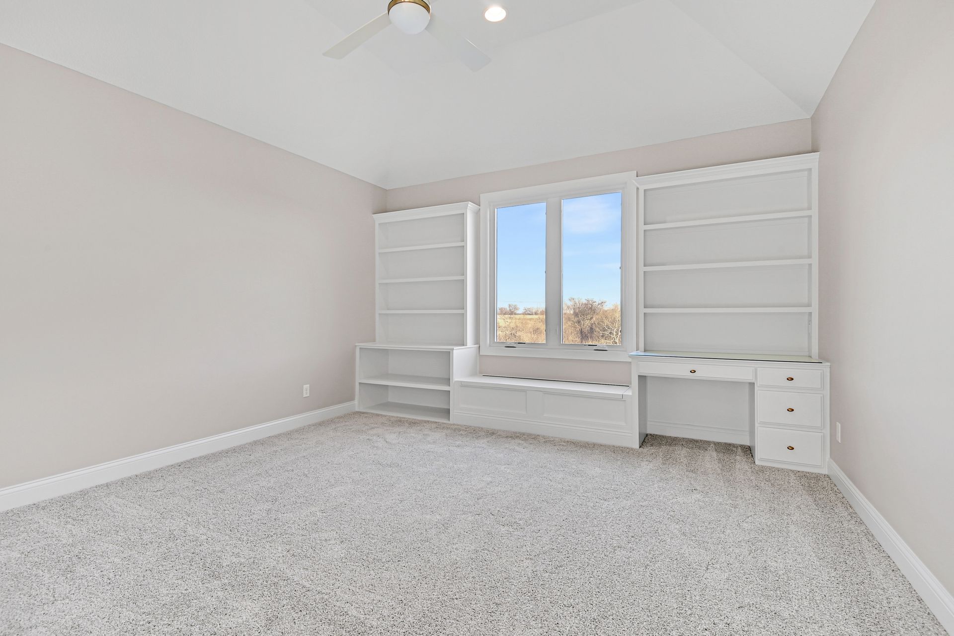 An empty room with a window , shelves , a desk and a ceiling fan.