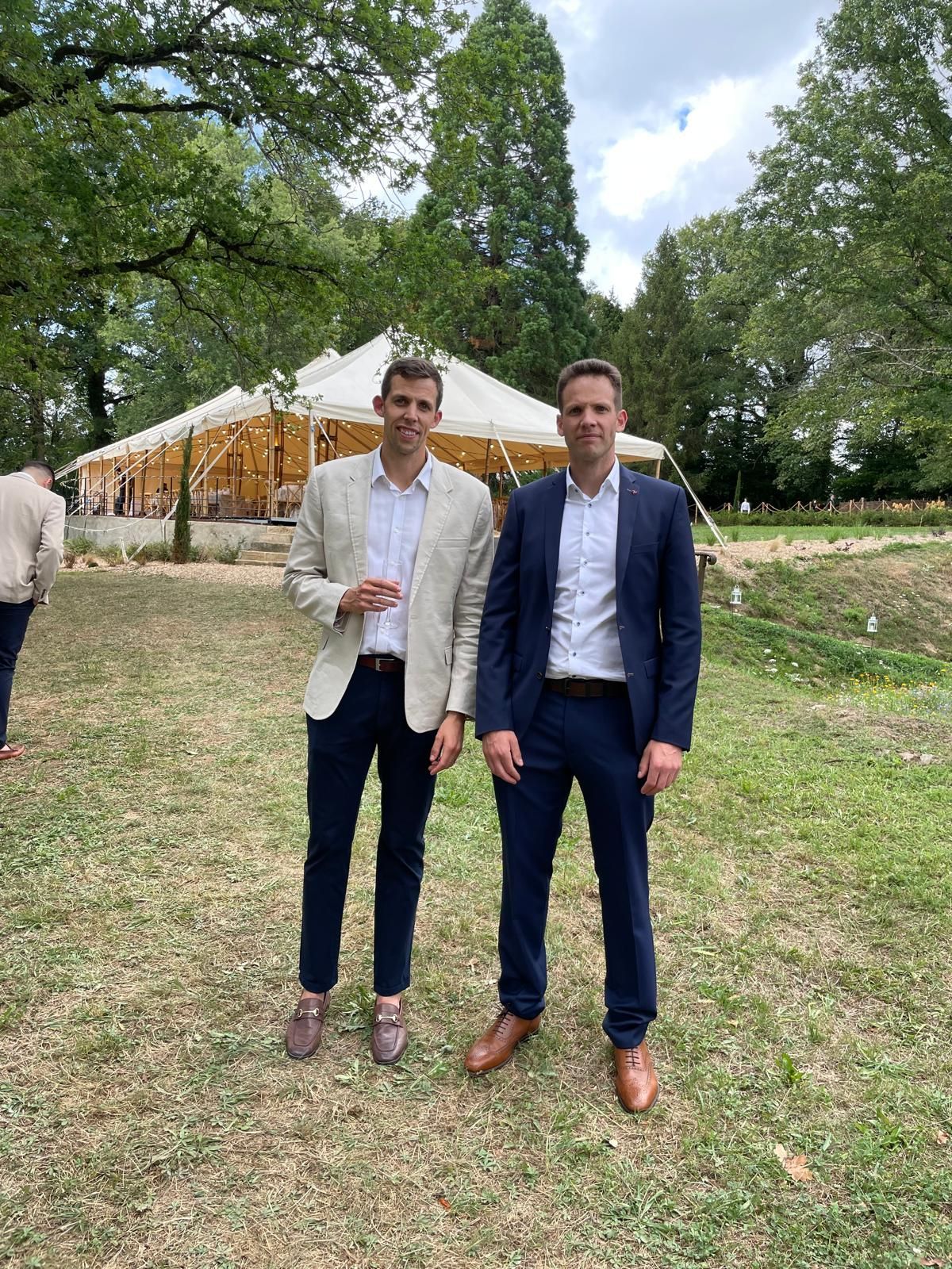 Two men in suits stand in a grassy area with a tent in the background.