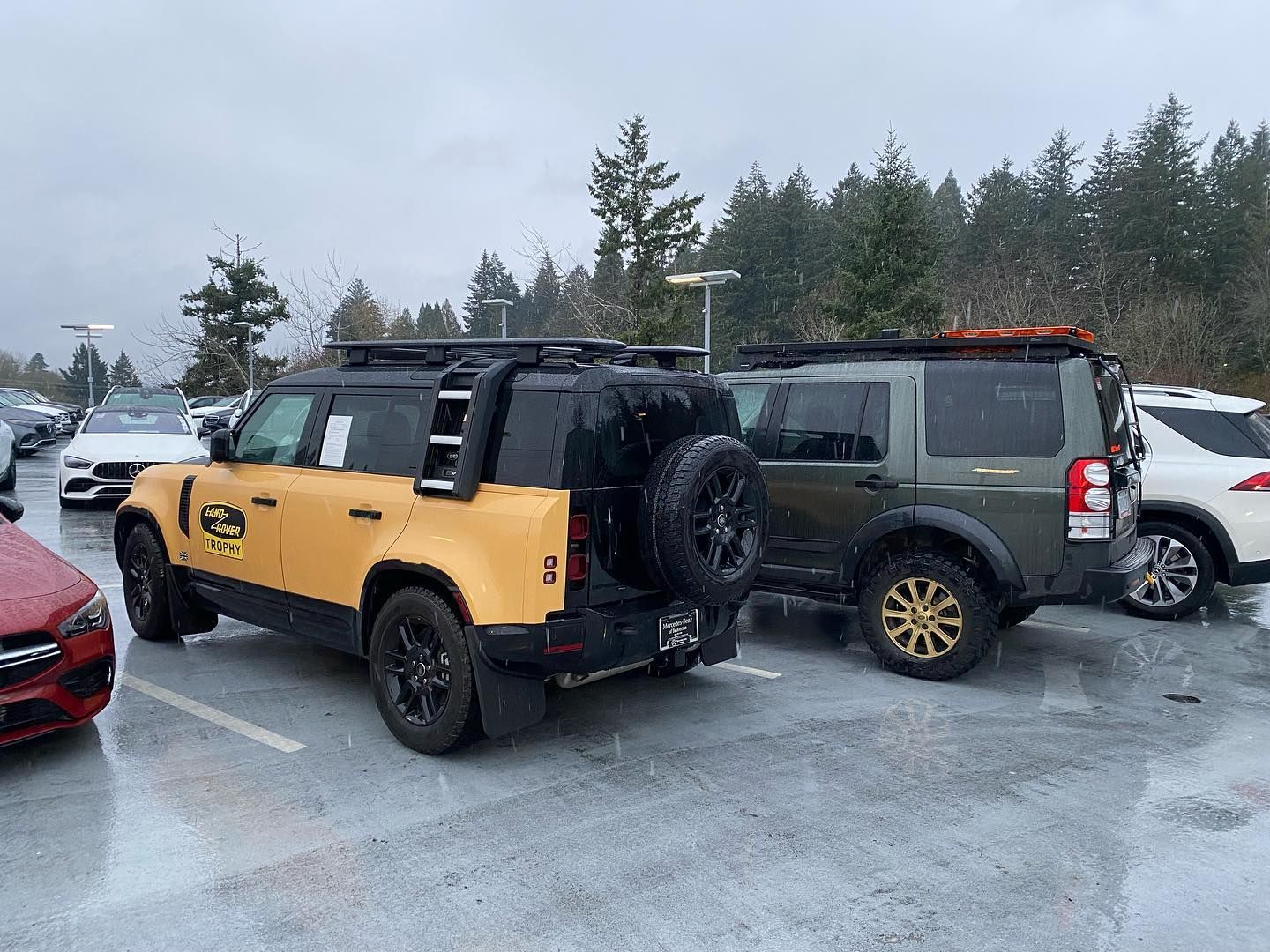 A yellow and black Land Rover Defender and an olive green Land Rover parked in a wet, snowy lot with a forest backdrop. | West Coast Overland