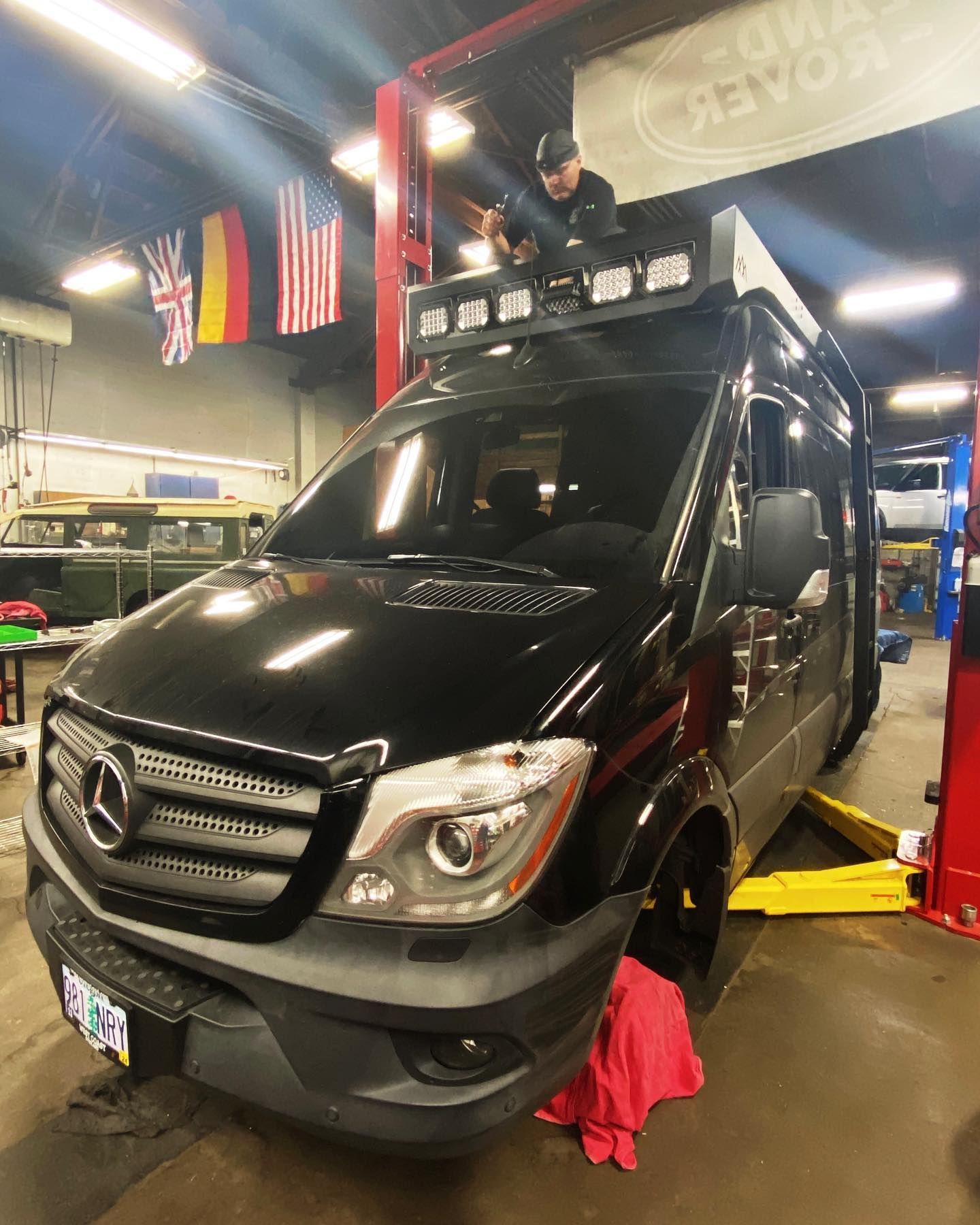 A person posing on the roof of a black Mercedes-Benz Sprinter van inside an automotive repair shop. | West Coast Overland