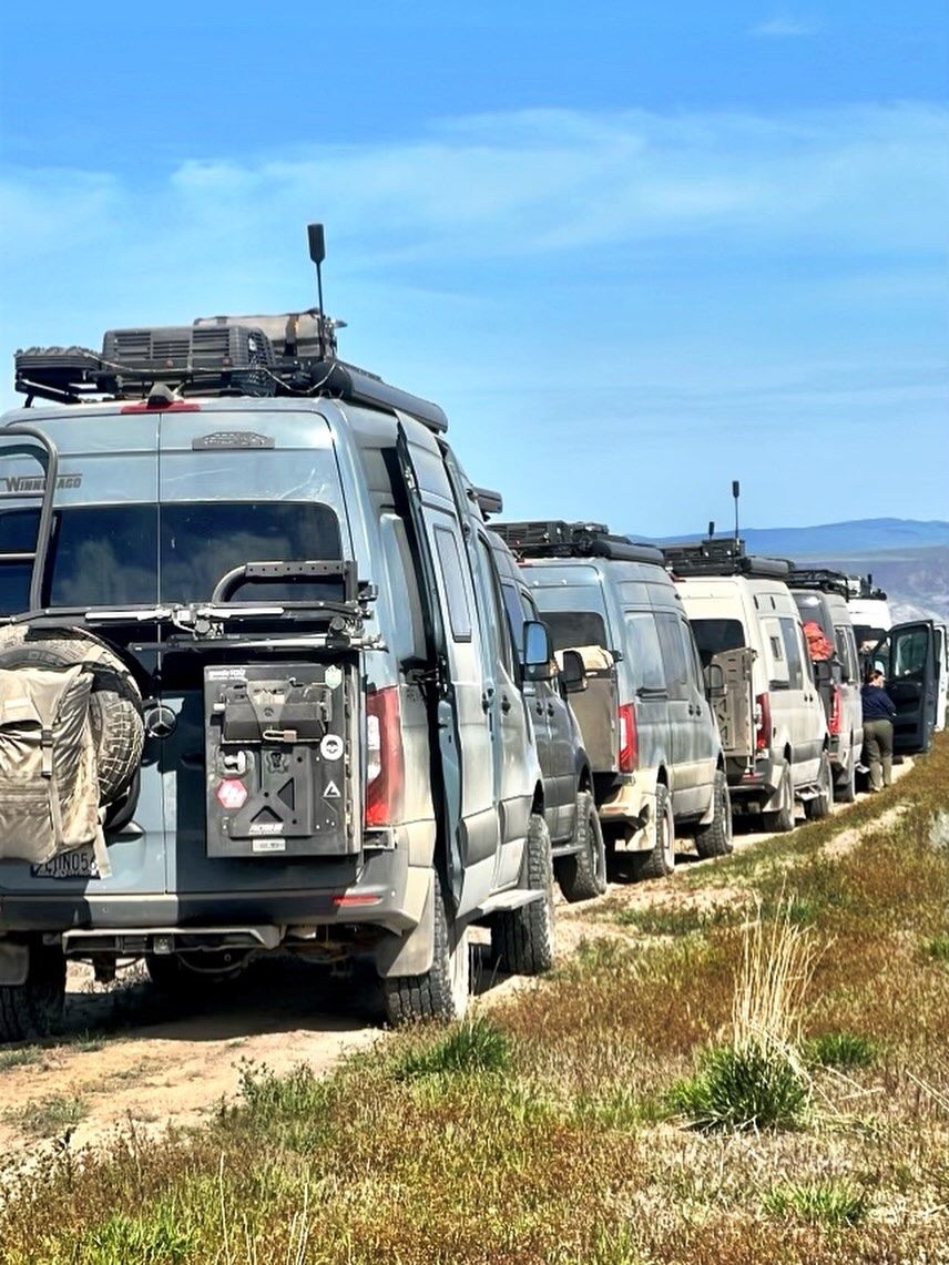 A caravan of gray adventure vans parked in a line on a dirt road under a blue sky. | West Coast Overland
