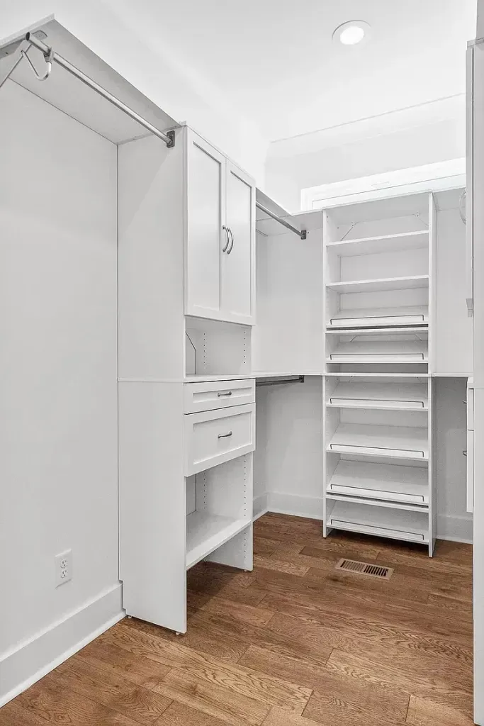 A walk in closet with white cabinets and shelves and a wooden floor.