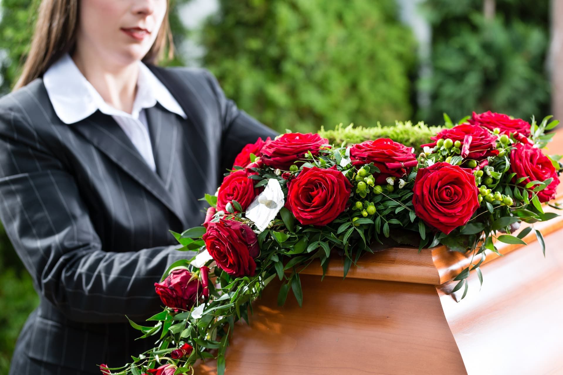 A professional in a suit standing by a wooden casket adorned with red roses and greenery.