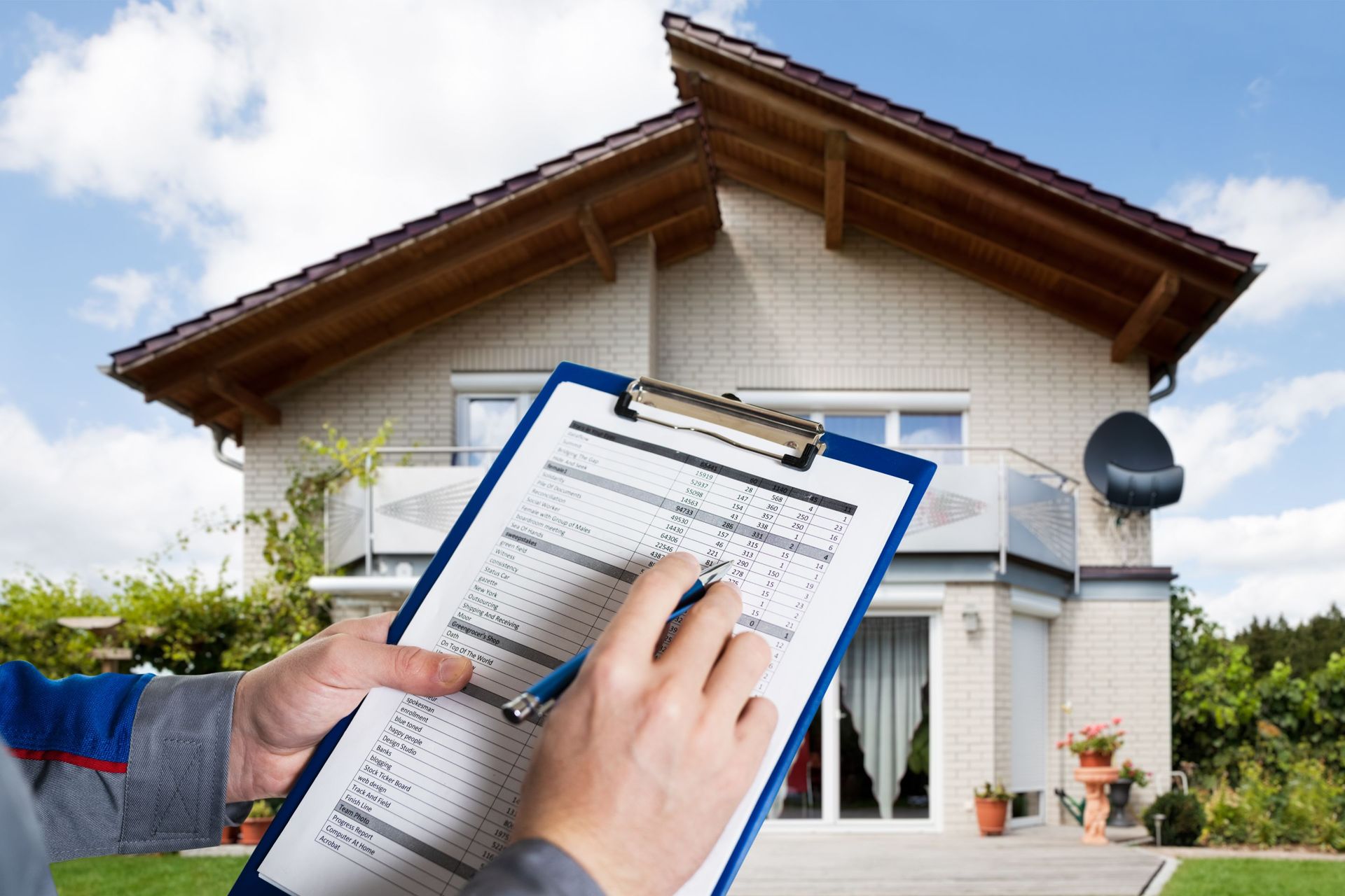 Person holding clipboard, inspecting a house exterior under a blue sky.