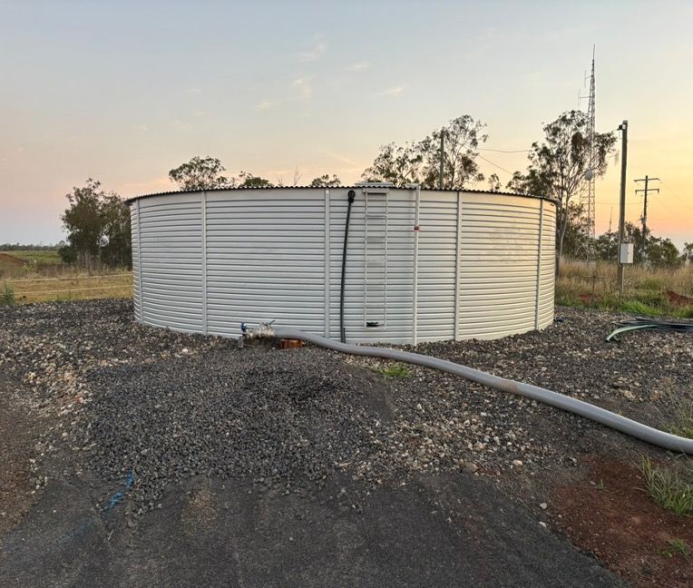 A large, circular corrugated steel water tank in a rural setting, with a hose connected. — Gin Gin Domestic Water Supplies in Wallaville, QLD