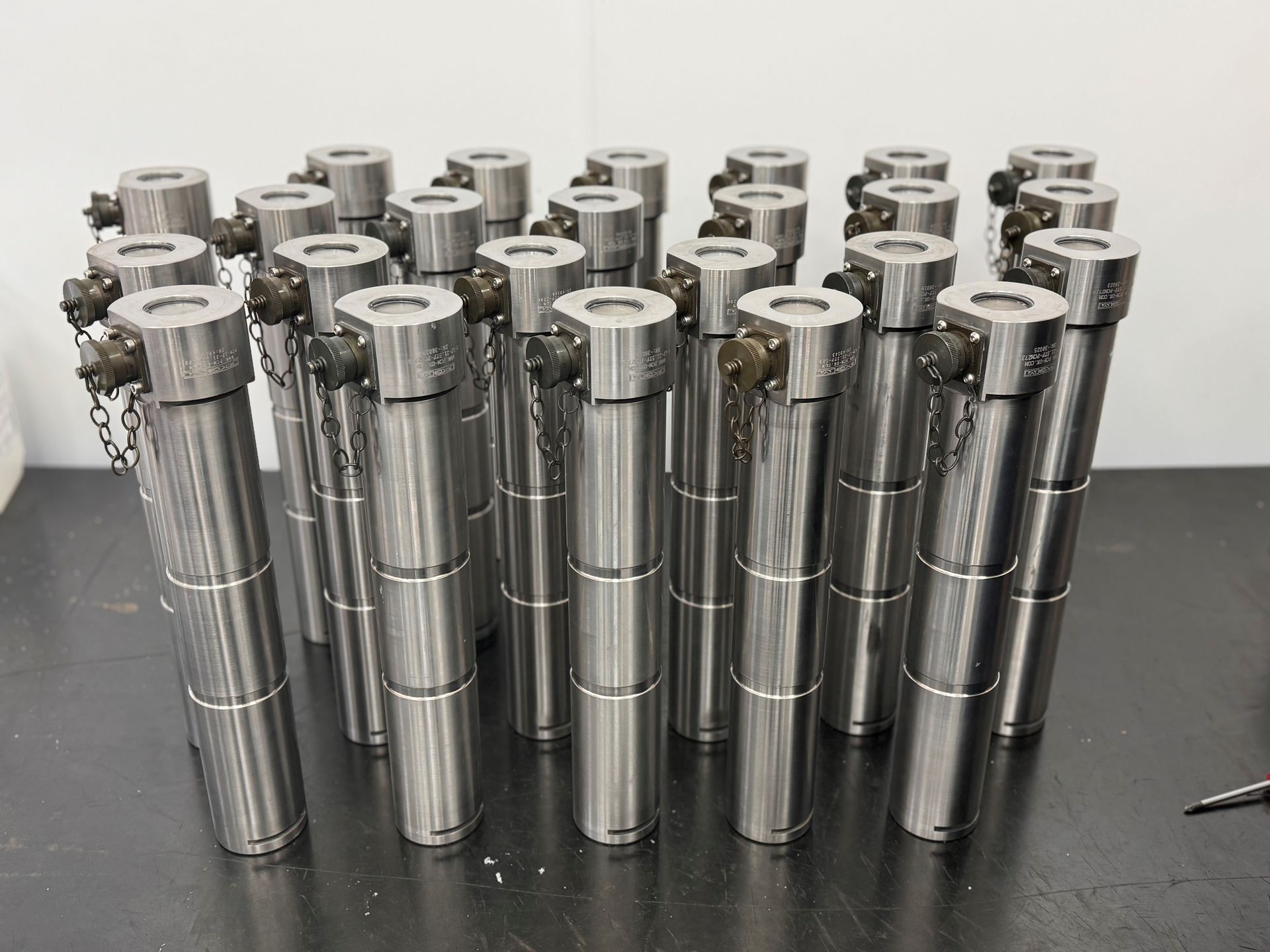 A large batch of stainless steel load pins from Procter and Chester, laid out on a black tabletop with white walls.