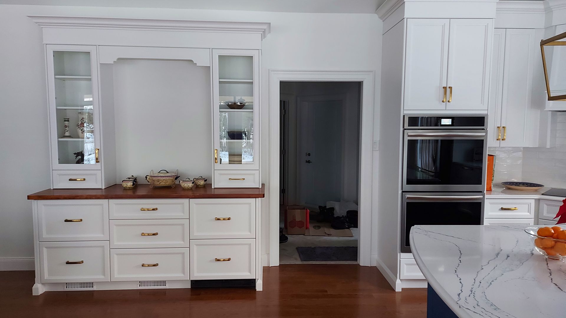 White kitchen with built-in cabinets, oven, and countertop with fruit; an open door leads to another room.