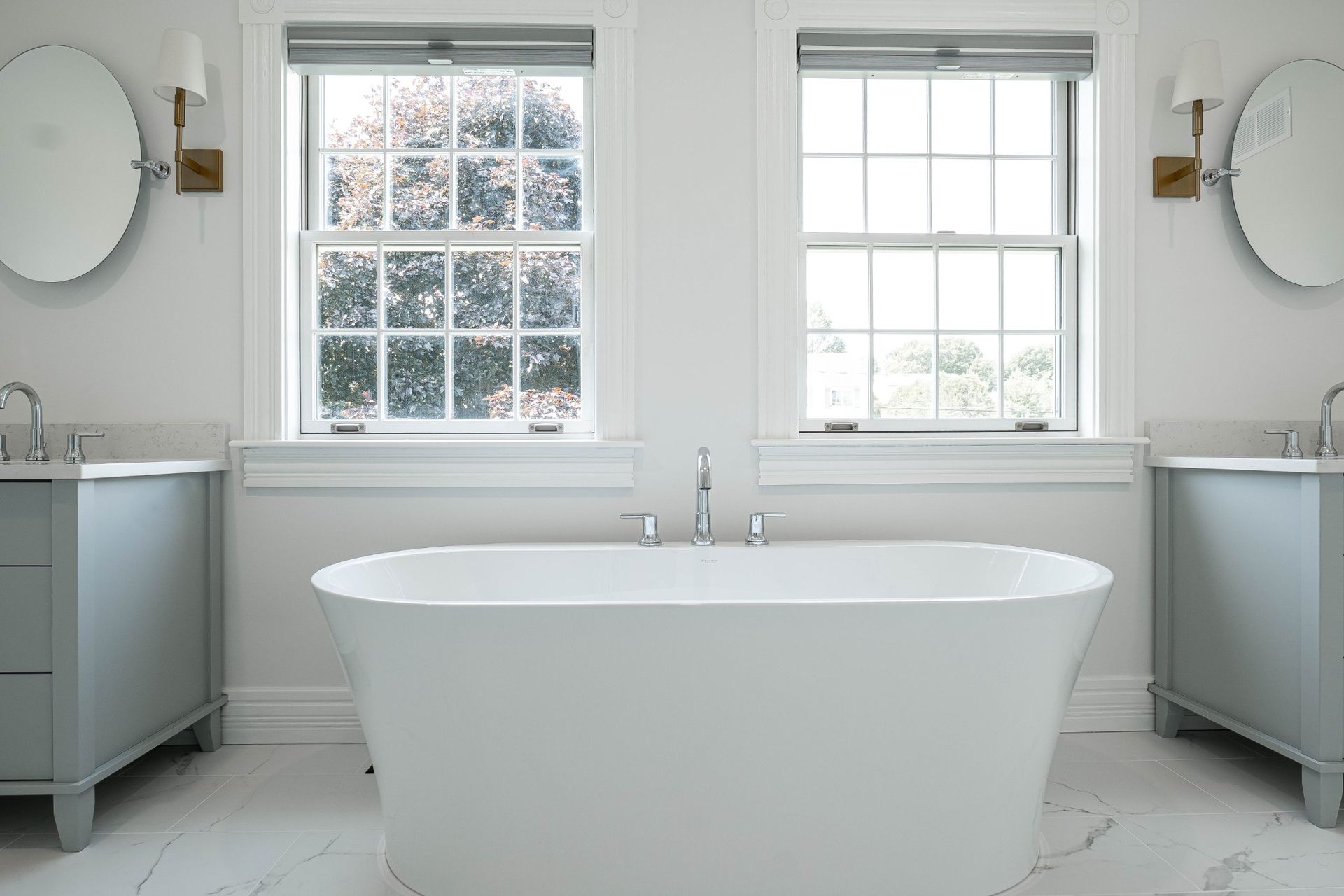 Bathroom with a white bathtub, gray cabinets, two windows, and round mirrors.