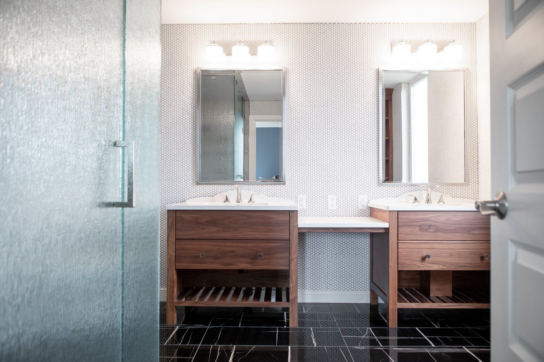 Modern bathroom with two wooden vanities, mirrors, and patterned wallpaper. Black tiled floor, glass shower door.