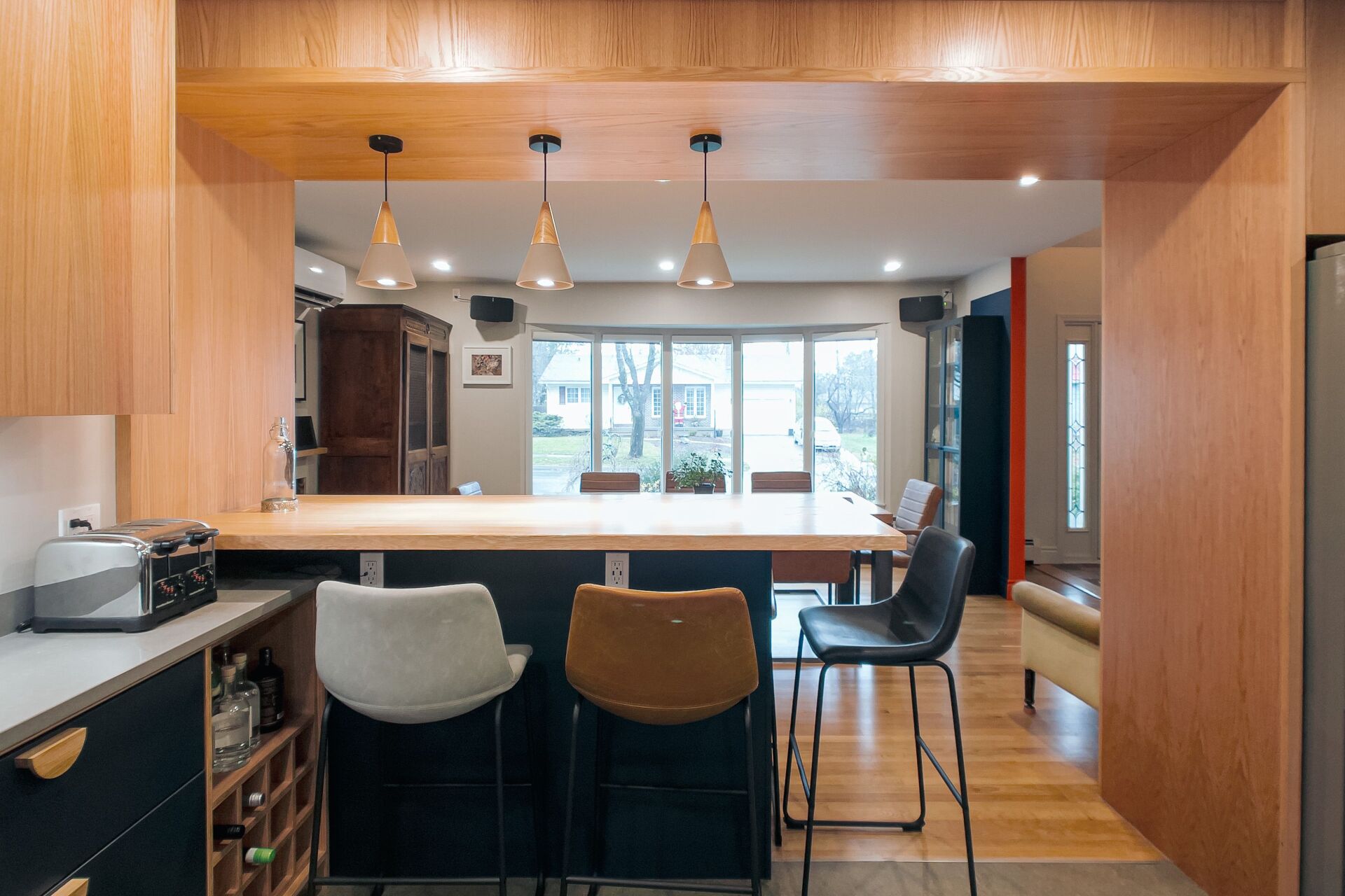 Kitchen with a bar and stools. Wood cabinets, three hanging lights, and a view of a dining area.