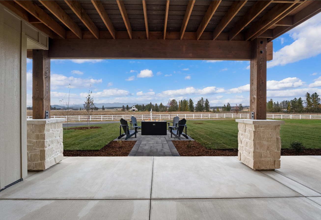A patio with a grill and chairs under a pergola with a view of a field.