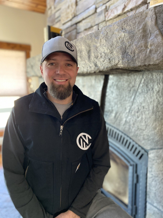 A man wearing a black vest and a hat is sitting in front of a fireplace.