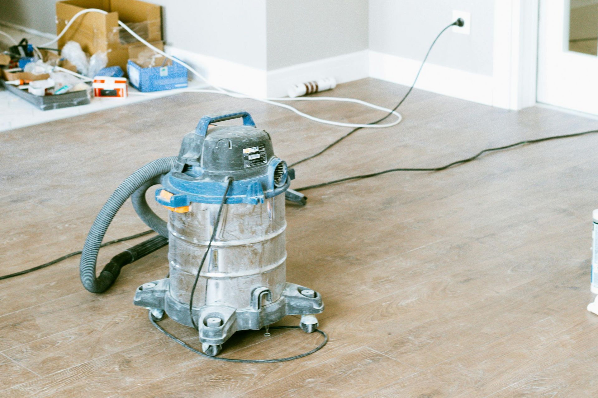 Wet/dry vacuum on a light brown floor in a room, with electrical cords, boxes, and a white baseboard visible.