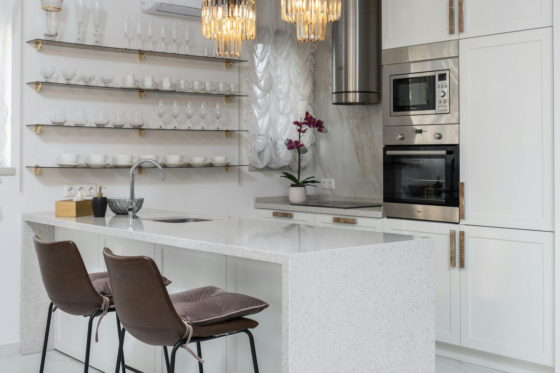 Modern white kitchen with island, seating, and built-in appliances; shelves with glassware.