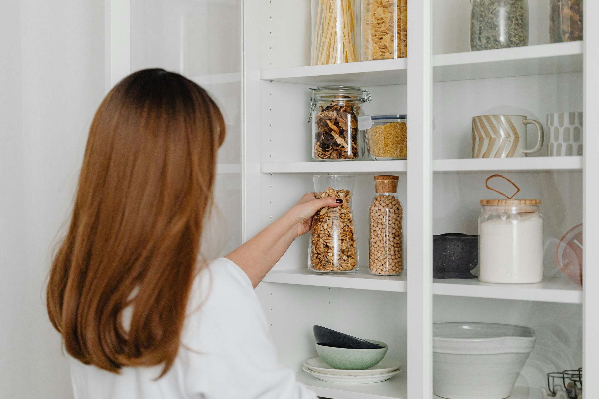 Woman in white shirt reaching for a jar of food in a white pantry.