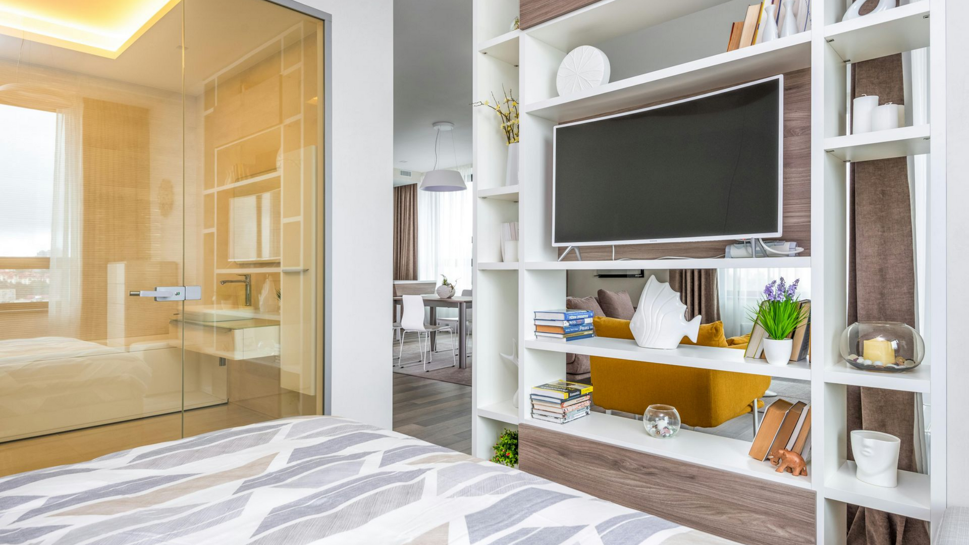 Bedroom with TV built into white and wood-tone shelving; bathroom visible through glass wall.