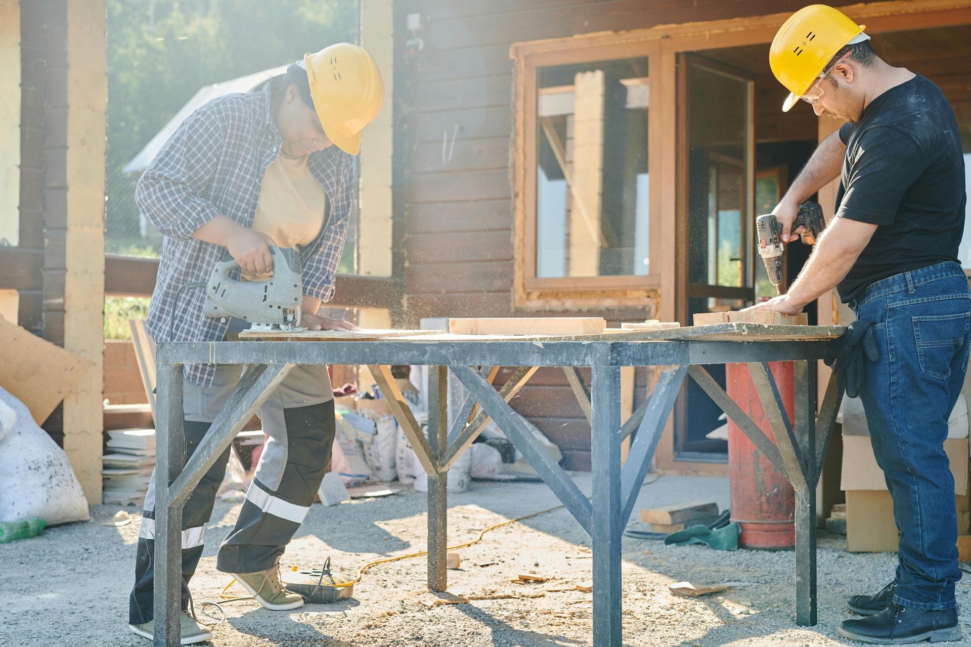 Two people in yellow hard hats working with power tools at a wood table outdoors.