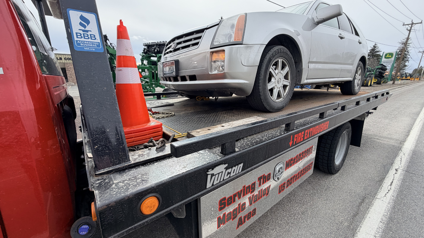 A silver Cadillac SUV is secured on the bed of a red flatbed tow truck with an orange safety cone nearby.