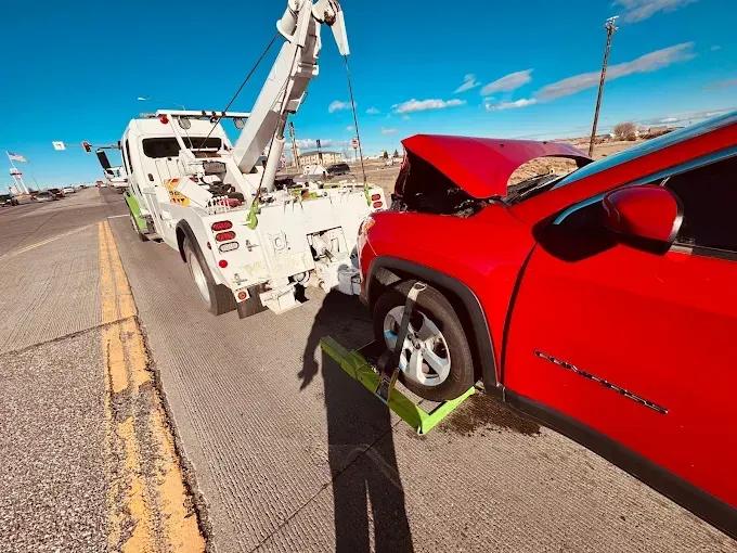 A red car with an open hood is hooked to a white tow truck on the side of a road under a clear blue sky.