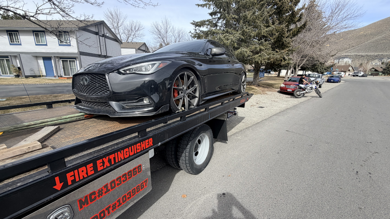 A dark grey Infiniti coupe secured on the bed of a flatbed tow truck parked along a residential street.