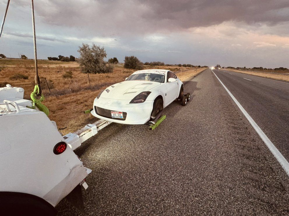 A white sports car being towed along the shoulder of a rural highway at sunset.