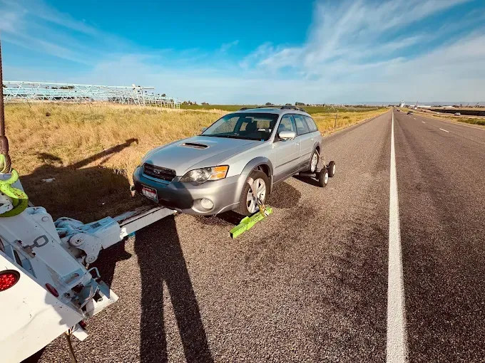 A silver Subaru station wagon being towed by a wrecker on the shoulder of a highway under a clear blue sky.