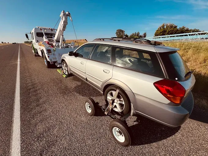A silver station wagon is being towed on a highway using a tow dolly under its rear tires by a white tow truck.