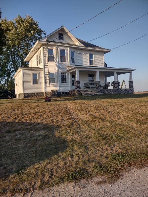 A large white house with a porch is sitting in the middle of a field.