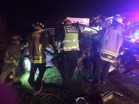 A group of firefighters are standing around a wrecked car at night.