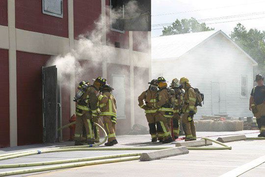 A group of firefighters are standing in front of a building with smoke coming out of it during a training exercise.