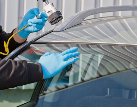 A person wearing blue gloves uses a professional tool to apply sealant to the edge of a car windshield.