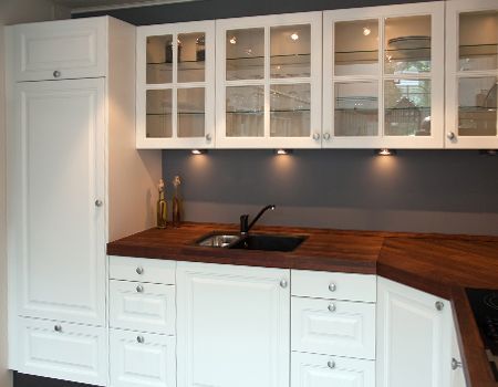 A white kitchen with a dark wood countertop, black sink, glass-front upper cabinets, and under-cabinet lighting.
