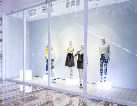 Four mannequins wearing stylish clothing stand in a brightly lit, minimalist retail window display.