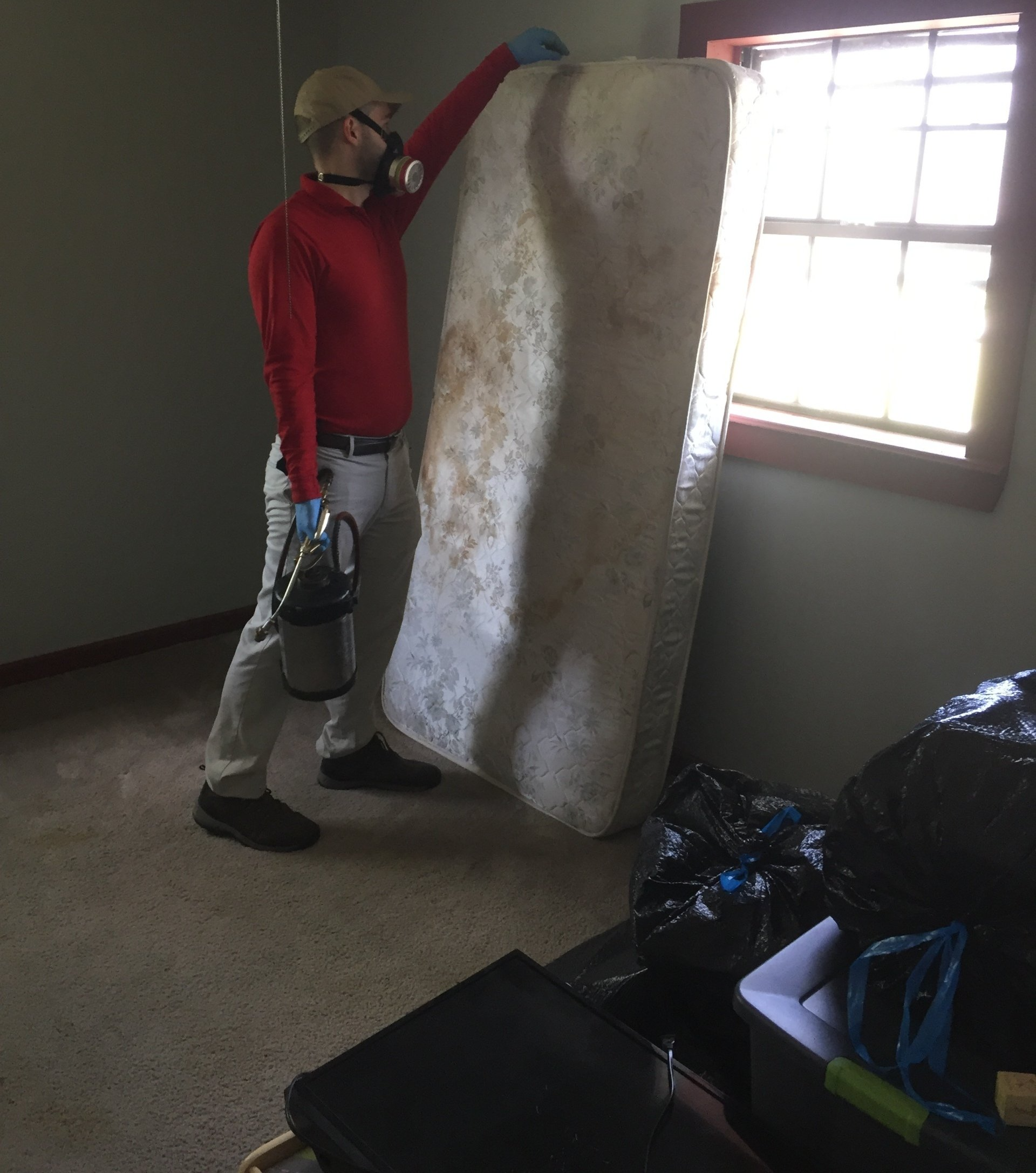 Person in protective gear holding a stained mattress in a room with a window; trash bags are visible.