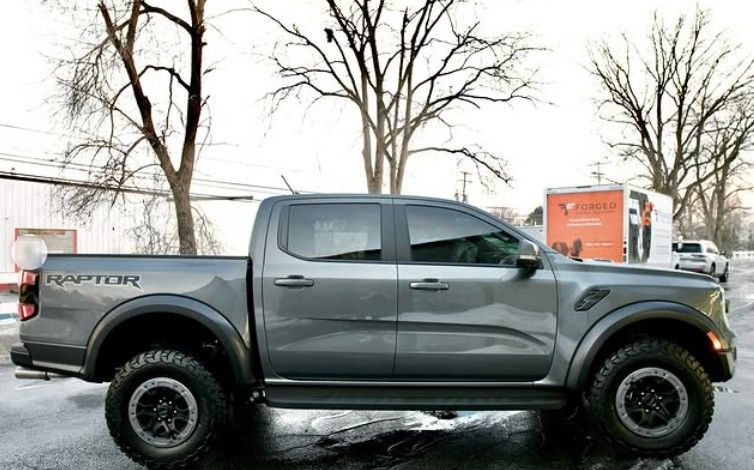 A gray Ford Ranger Raptor pickup truck parked outdoors against a backdrop of bare trees and a white trailer.